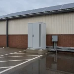 Freestanding commercial ESS battery storage cabinet beside a factory wall with switchgear and rooftop solar, the utility-scale scope behind ESS battery cost per kWh
